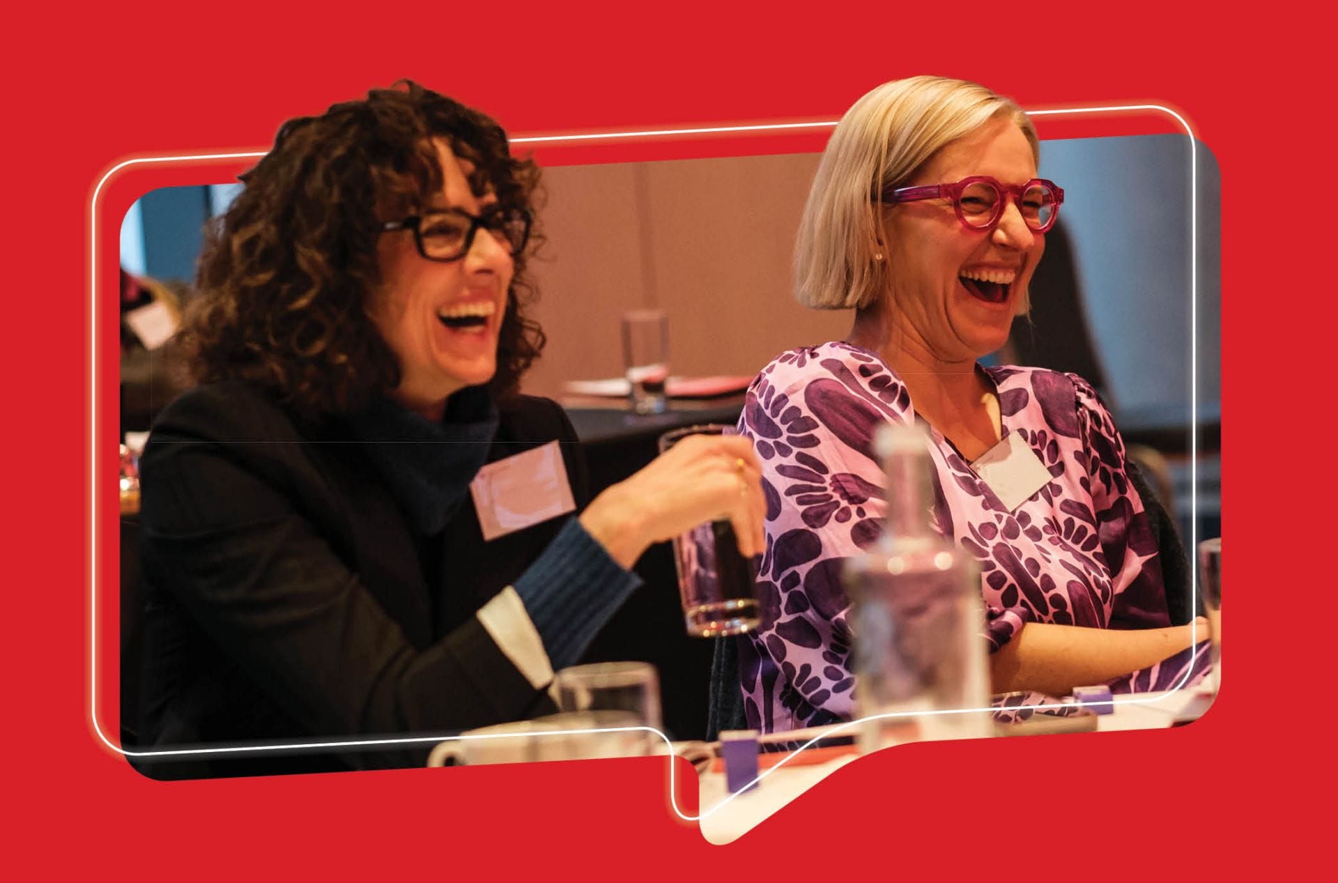 Two women sitting at a table at the Women's leadership symposium looking to the right and laughing
