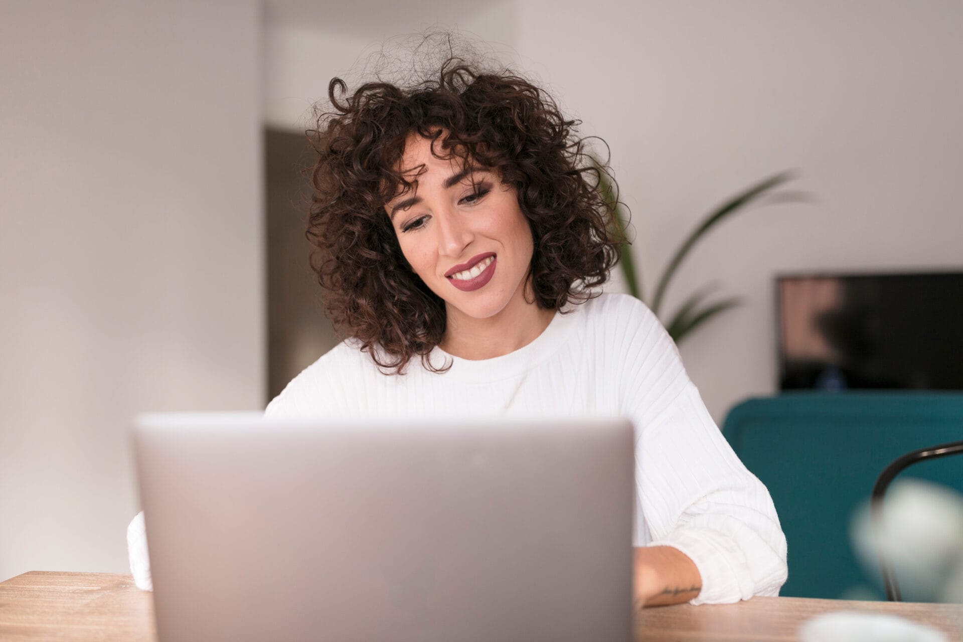 A woman with dark curly hair sits at her laptop.