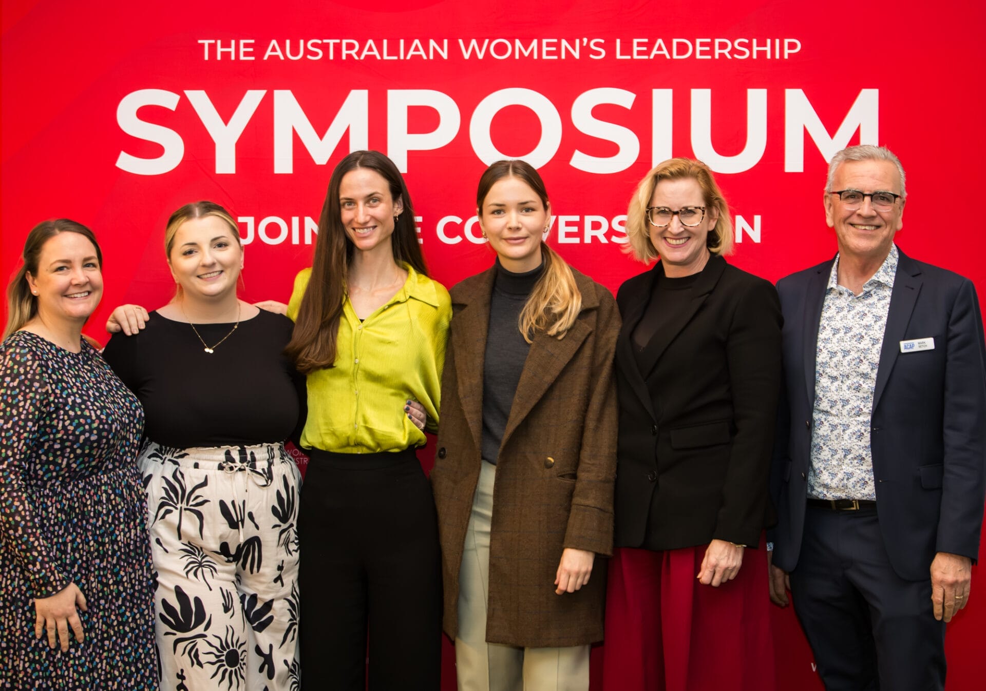 A happy group of women and man stand infront of a red banner with the words The Australian Women's Leadership Symposium