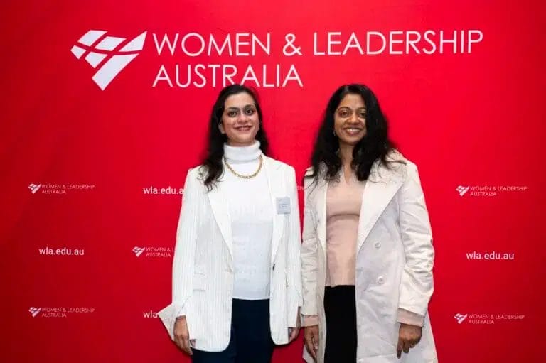 Symposium. Two women wearing white jackets stand infront of a media wall that says Women & Leadership Australia.